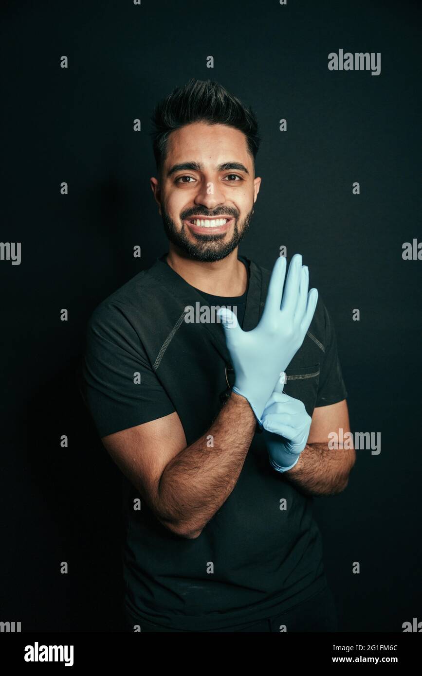 Porträt eines Mannes mit gemischter Rasse, der OP-Handschuhe trägt und mit glänzenden Zähnen vor schwarzem Hintergrund lächelt Stockfoto