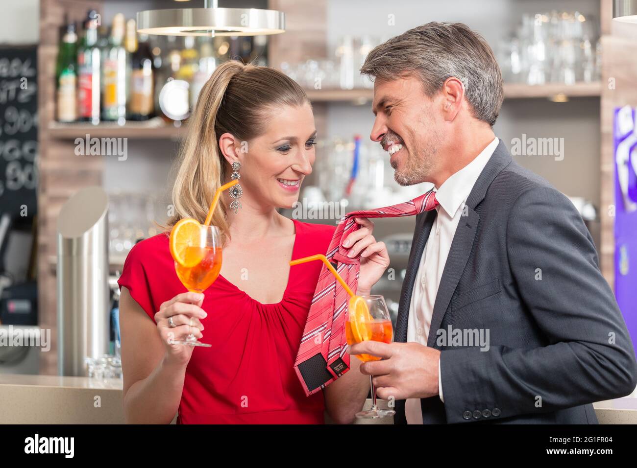 Lächelnde Frau hält ihre Ehemann Krawatte, während sie einen orangenen Cocktail in einer anderen Hand hält Stockfoto