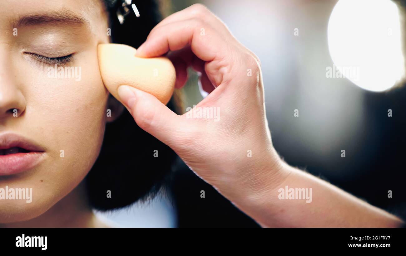 Teilansicht eines Make-up-Künstlers mit kosmetischem Schwamm, der Gesichtsfundament auf ein Modell mit geschlossenem Auge aufgibt Stockfoto