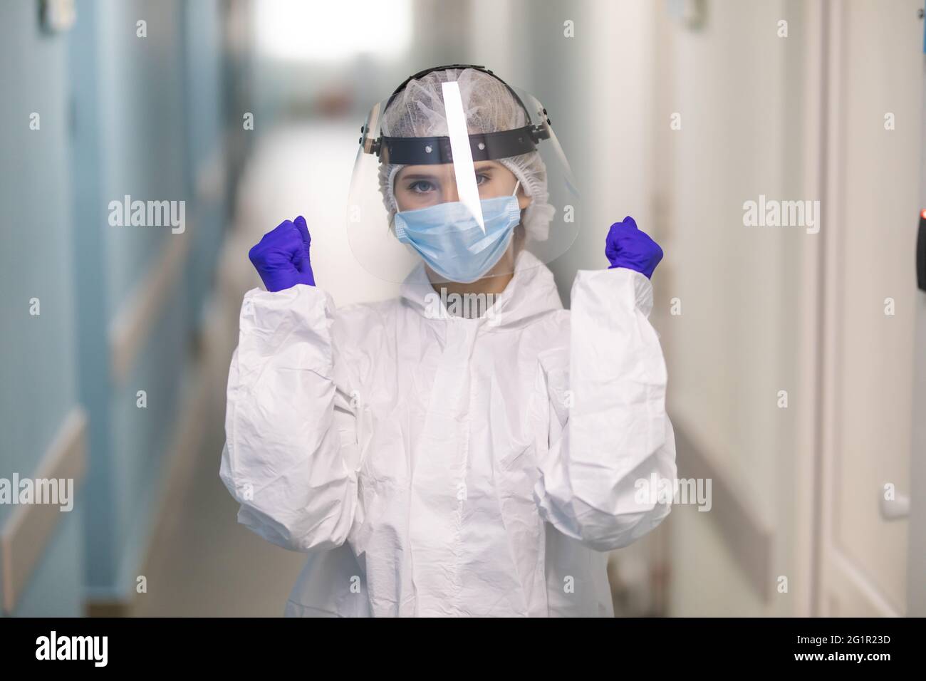 Porträt einer jungen Krankenschwester mit einer WIN-Geste mit einem Gesichtsschutz und einer medizinischen Maske, die am Krankenhausflur steht. Coronavirus-Prävention Stockfoto