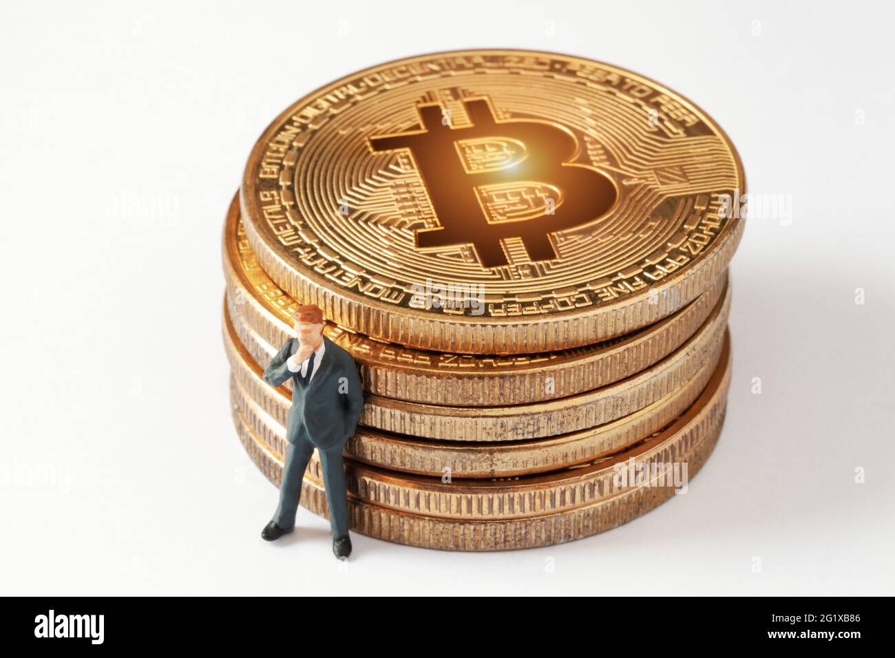 Winzig denkende Geschäftsmann Figur, während sie sich zu glänzendem Bitcoin Stapel auf weißem Hintergrund lehnt. Entwicklung einer Anlagestrategie für Kryptowährungen. Stockfoto