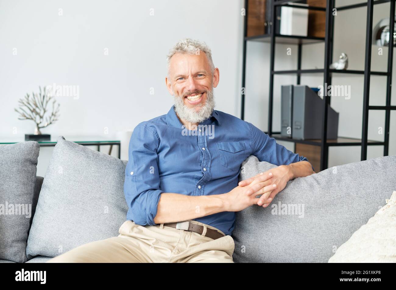 Ein gutaussehender, optimistischer, reifer Mann, der mit einem fröhlichen, toothy Lächeln auf die Kamera starrt, ein glücklicher älterer Mann in eleganter Freizeitkleidung, der zu Hause auf dem Sofa sitzt. Lifestyle- und Wohlfühlkonzept Stockfoto