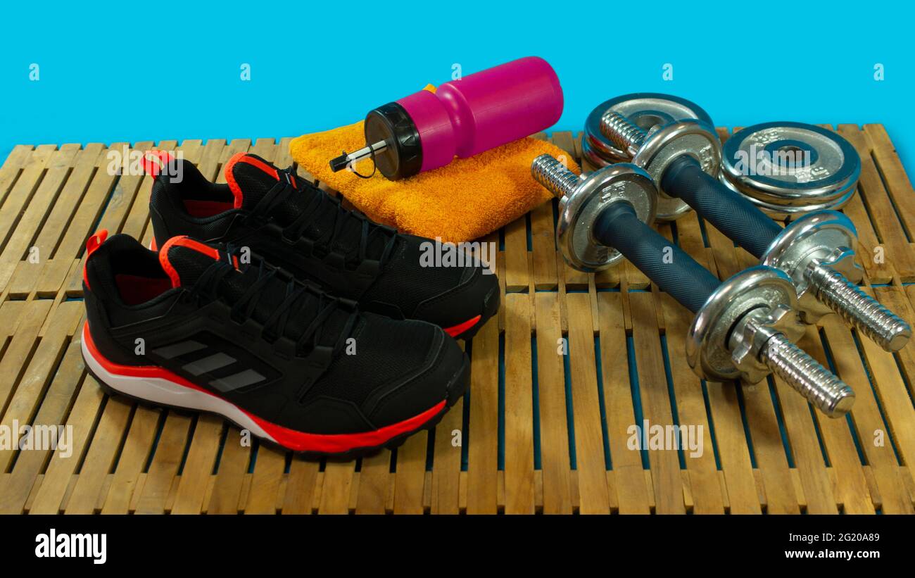 Gym-Set aus schwarzen Sportschuhen, Hanteln zum Trainieren, gelbem Handtuch und roter Flasche mit Wasser auf Holzboden mit blauem Hintergrund Stockfoto