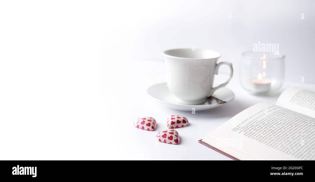 Wunderschöne weiße, entspannende Leseszene mit offenem Buch, Tasse, Kerze und herzförmigen Schokoladenbonbons. Stockfoto