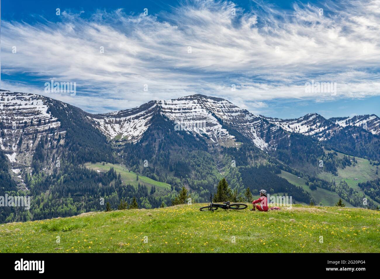 Lächelnde ältere Frau, die mit ihrem elektrischen Mountainbike unter den schneebedeckten Bergen der Nagelfluh-Kette bei Oberstaufen, Allgäu, Bayern, fährt Stockfoto