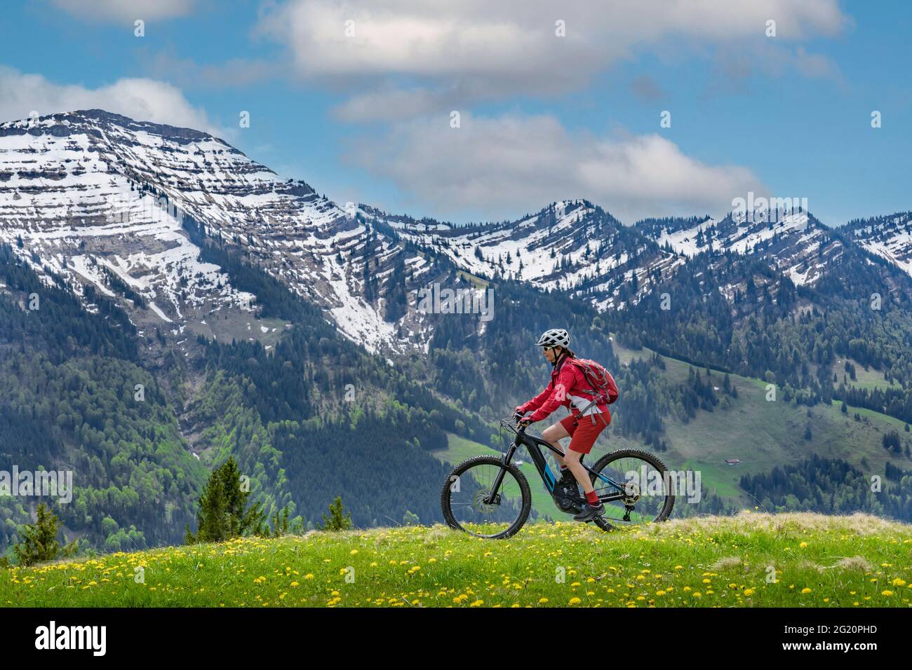 Lächelnde ältere Frau, die mit ihrem elektrischen Mountainbike unter den schneebedeckten Bergen der Nagelfluh-Kette bei Oberstaufen, Allgäu, Bayern, fährt Stockfoto