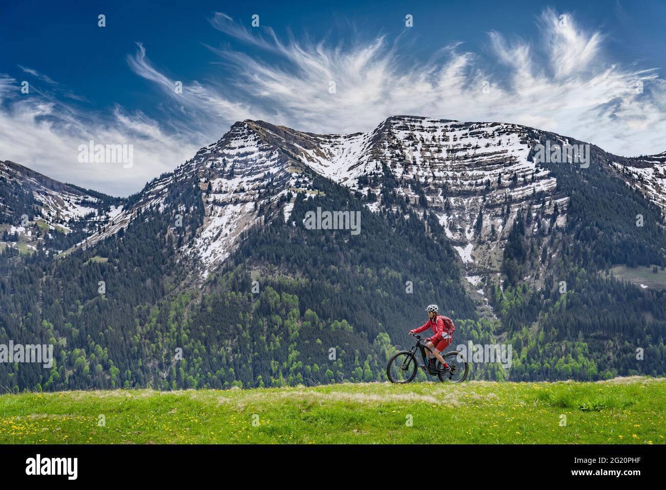 Lächelnde ältere Frau, die mit ihrem elektrischen Mountainbike unter den schneebedeckten Bergen der Nagelfluh-Kette bei Oberstaufen, Allgäu, Bayern, fährt Stockfoto