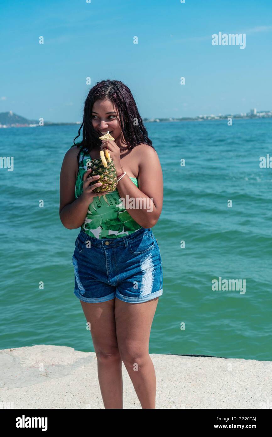 Ziemlich gebräunte Frau, in Shorts gekleidet, trinkt einen Cocktail am Strand. Konzept von Reisen und Entspannung. Helle Farben und sonniger Tag. Stockfoto