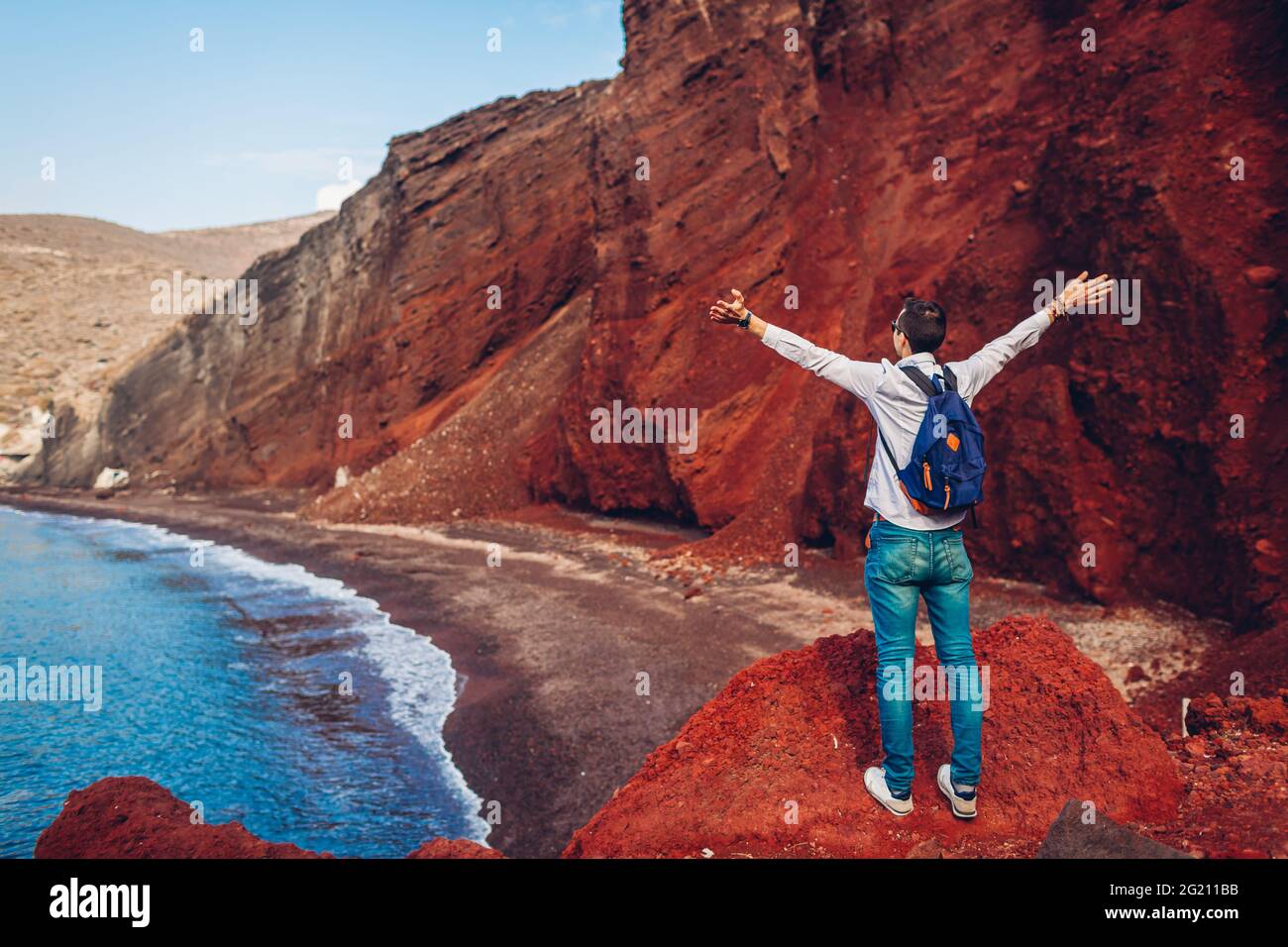 Reisende, die die rote Strandlandschaft mit aufgehobenen Armen betrachten, fühlen sich glücklich in Akrotiri, Insel Santorini, Griechenland. Mann, der während der Herbstferien reist Stockfoto