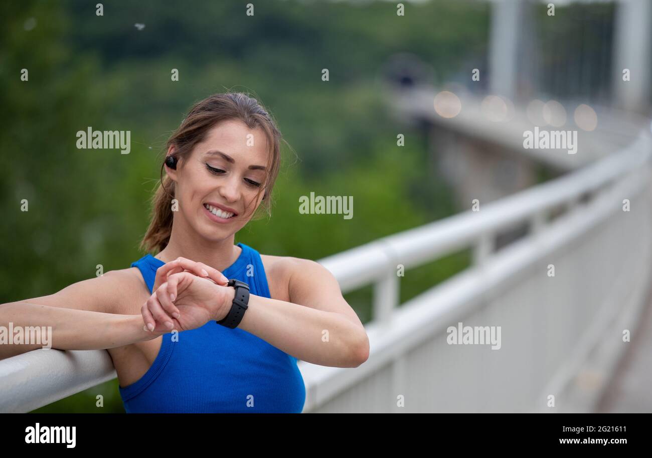 Weibliche Athletin, die eine intelligente Uhr zum Laufen aufsetzt. Junge athletische Frau, die sich beim Training auf dem Brückengeländer stützt. Stockfoto