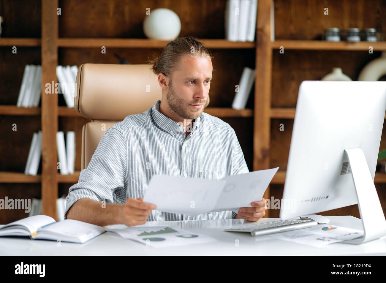 Ein kluger, junger Kaukasier, der in stilvoller Kleidung am Schreibtisch sitzt, einen Computer benutzt, Dokumente und Diagramme durchschaut, über die Finanzstrategie nachdenkt und konzentriert arbeitet Stockfoto