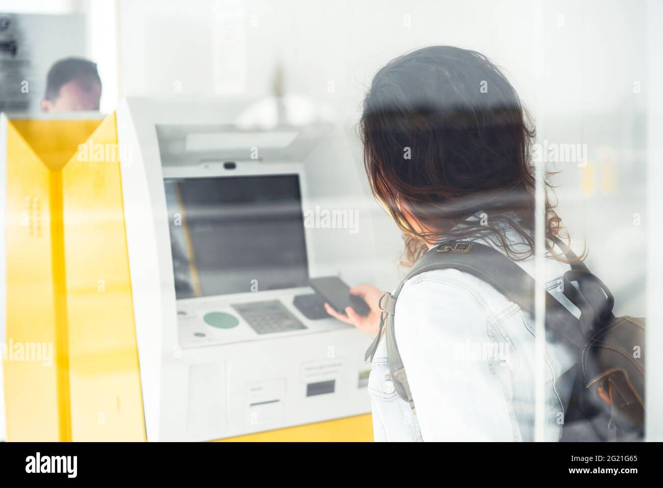 Hübsche junge Frau, die eine Schutzmaske trägt und am gelben Geldautomaten mit dem Telefon bezahlt. Moderne Banktechnologie hilft Touristen bei einfachen Zahlungen Stockfoto