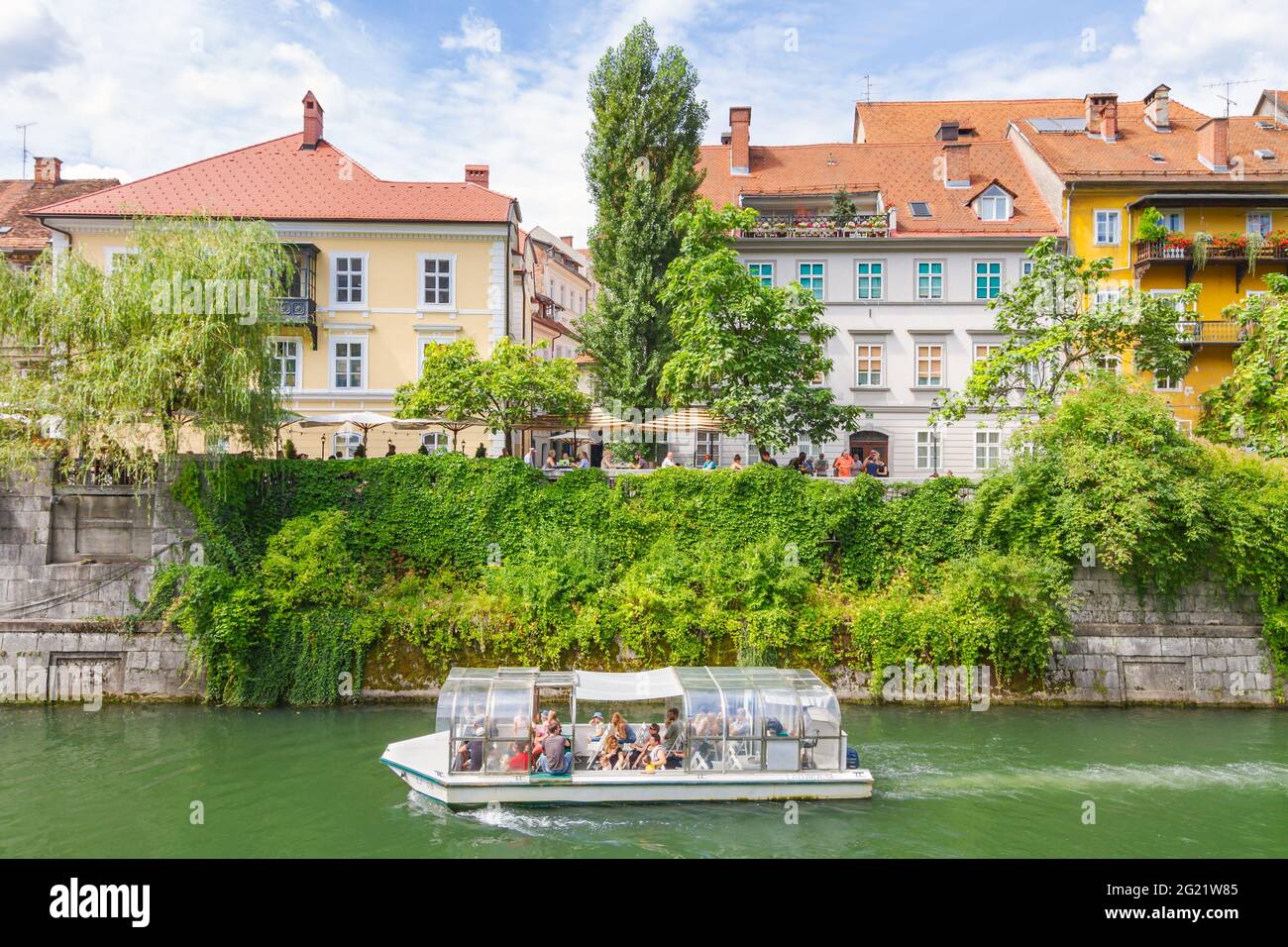 Ljubljana, Slowenien - 15. August 2018: Ein Touristenboot fährt auf dem Fluss Ljubljanica neben alten malerischen Gebäuden und grüner Vegetation in der Innenstadt vorbei Stockfoto