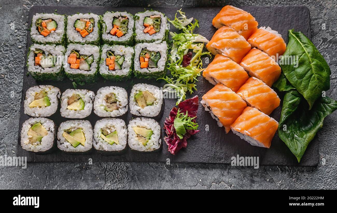 Lebensmittelbanner: Satz von verschiedenen Rollen auf einem Schneidebrett aus schwarzem Stein. Brötchen mit Lachs, Gemüse Sushi Maki-Brötchen und frischem Salat. Stockfoto