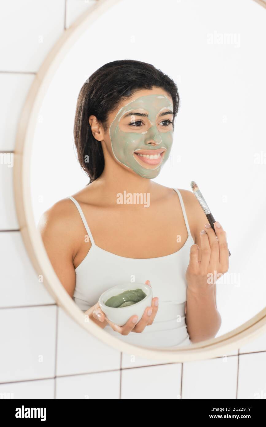 Lächelnde junge afroamerikanische Frau, die Maske mit Kosmetikbürste in der Nähe des Spiegels im Badezimmer aufführt Stockfoto