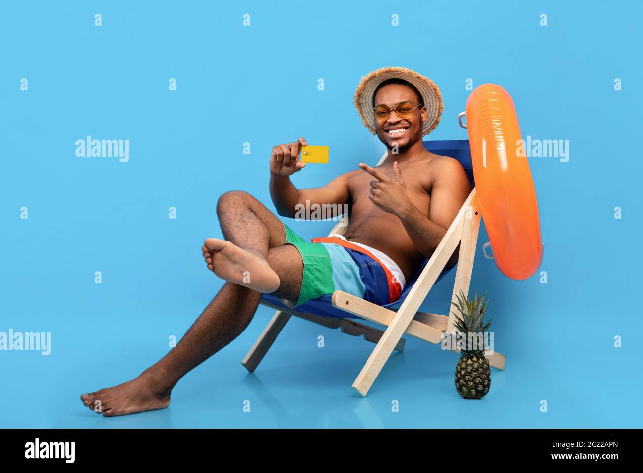 Fröhlicher schwarzer Kerl, der auf Kreditkarte zeigt, während er in den Sommerferien auf blauem Hintergrund im Liegestuhl sitzt Stockfoto