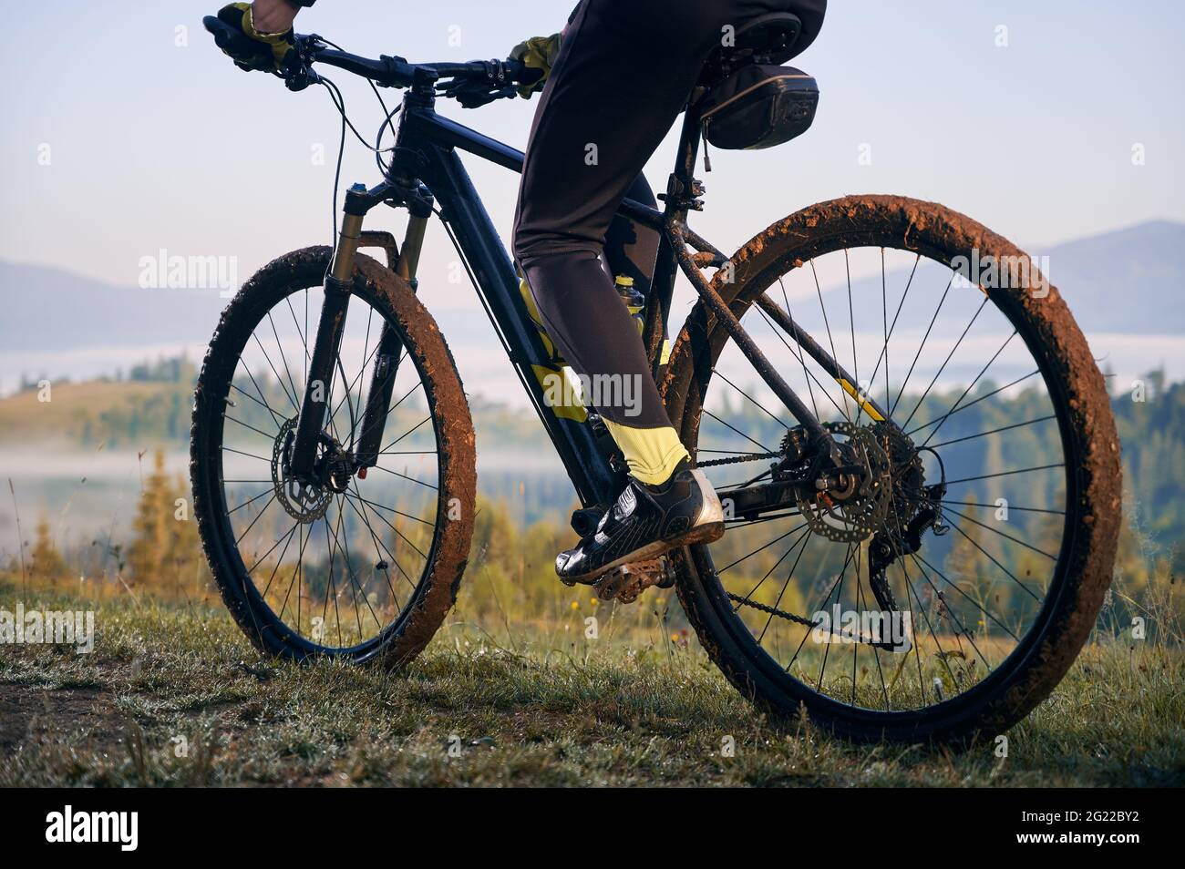 Horizontal geschnittene Momentaufnahme der Beine des Bikers, sitzend auf dem Fahrrad. Nahaufnahme auf dem Fahrrad, Räder im Schlamm, nach der morgendlichen Fahrt auf Bergwegen. Sportler, die den Moment des Sonnenaufgangs genießen Stockfoto