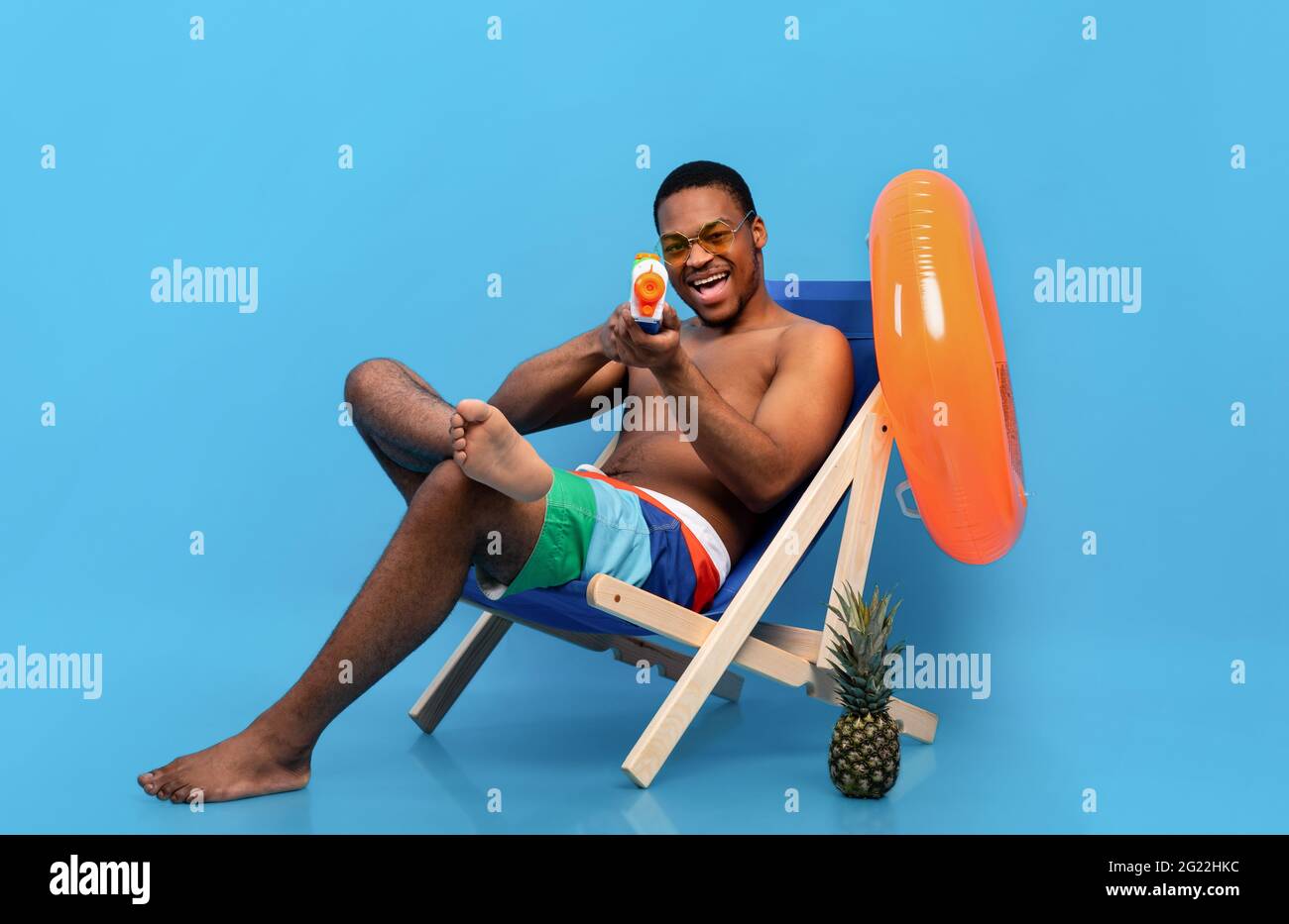 Verspielter schwarzer Mann, der sich mit einer Wasserpistole im Loungesessel entspannt und mit der Spielzeugpistole auf die Kamera über dem blauen Studiohintergrund zeigt Stockfoto