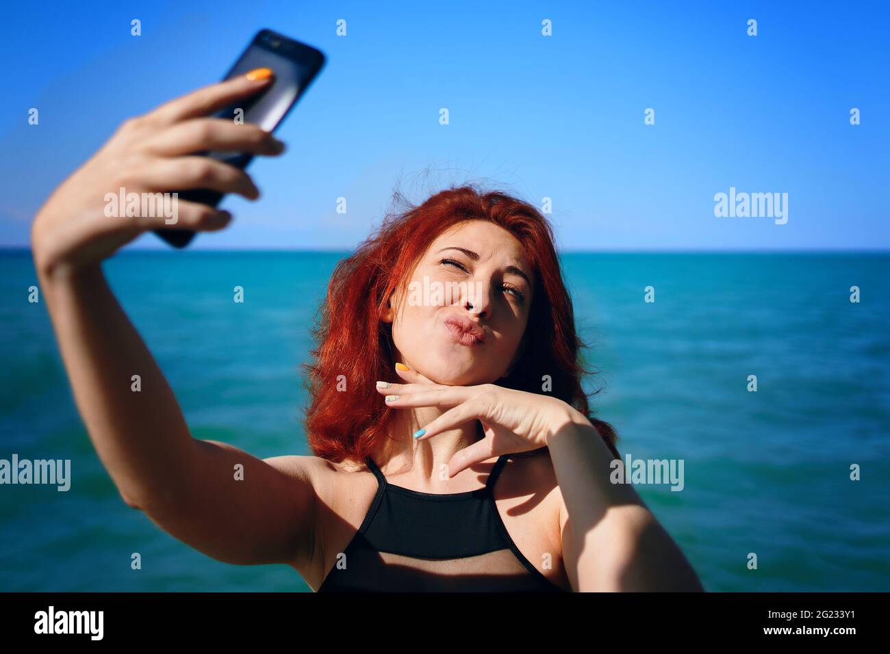 Rothaarige Frau schielt in der Sonne und macht Selfie auf der Smartphone-Kamera. Lippen wie eine Ente. Sonniger Sommertag. Nettes Mädchen macht Bilder von sich selbst vor dem Hintergrund des blauen Meeres und Himmel. Stockfoto