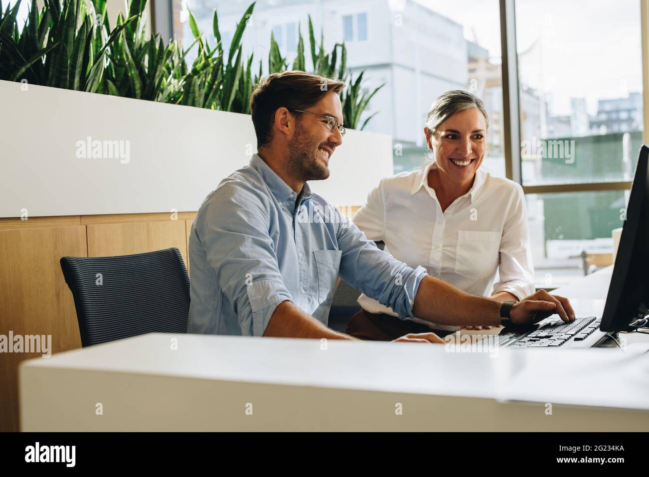 Manager hilft Mitarbeitern bei der Arbeit im Büro. Glückliche Frau mit mittlerem Erwachsenen, die mit einem jungen männlichen Kollegen zusammenarbeitet. Stockfoto
