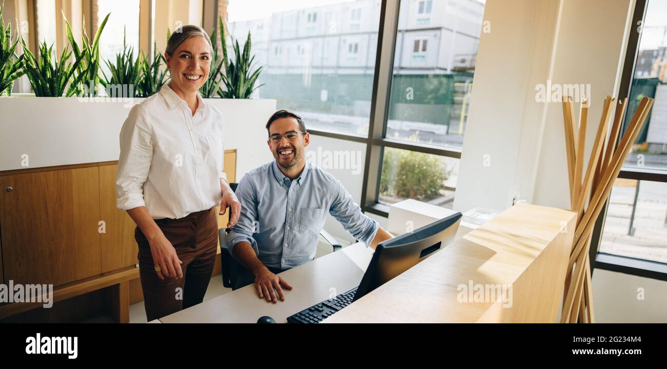 Reife Frau steht und junger Mann sitzt am Schreibtisch und lächelt in die Kamera. Führungskräfte, die in die Kamera schauen. Stockfoto