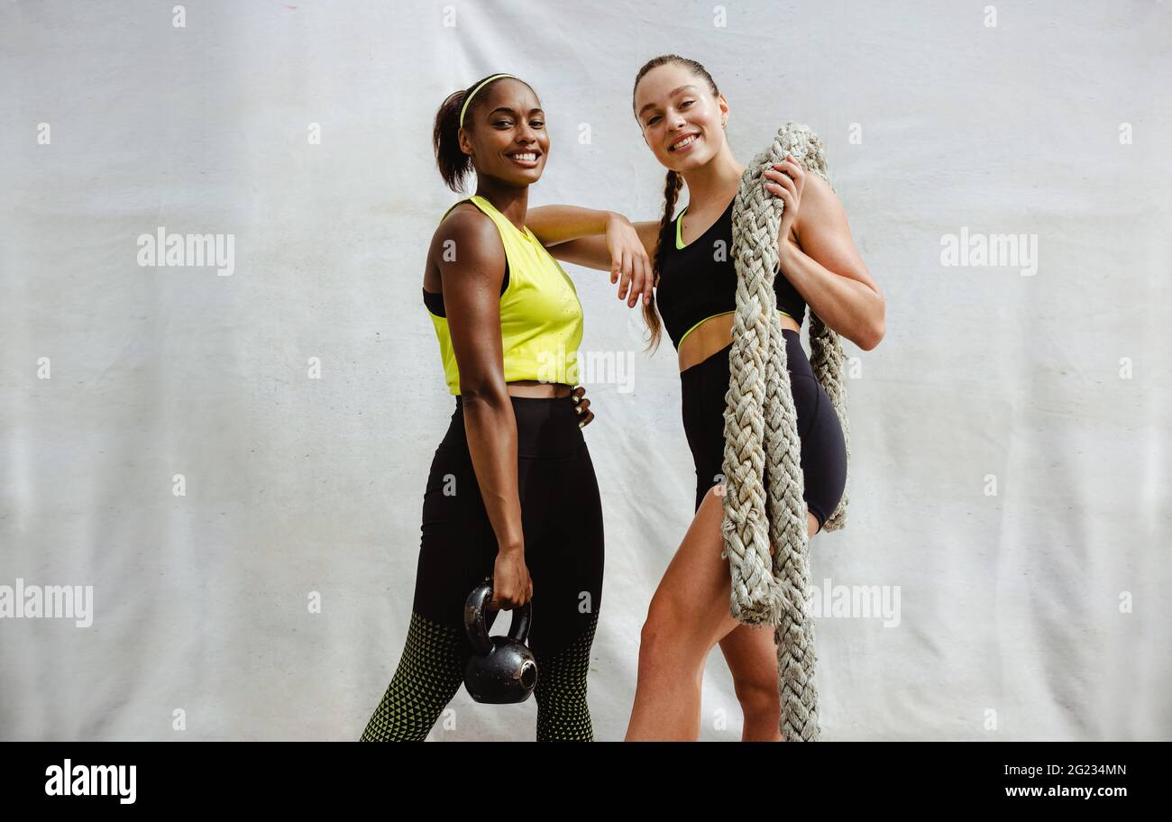 Fit Frau mit Kugelhantel und ihre Freundin hält Kampfseil. Frauen in Sportbekleidung stehen mit Fitnessgeräten auf weißem Hintergrund. Stockfoto