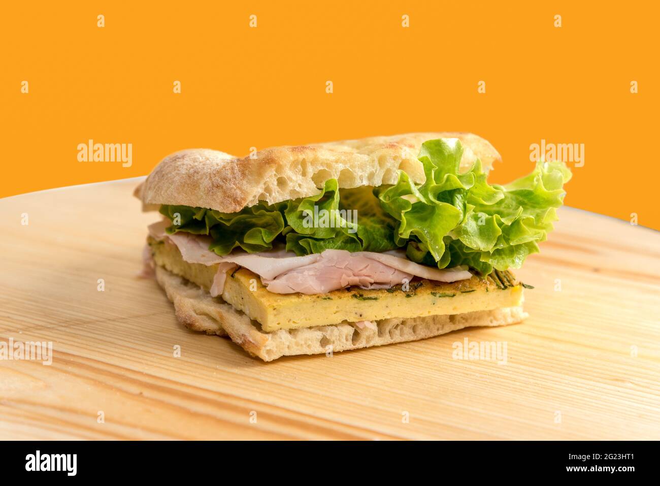Focaccia-Sandwich mit Salat, Schinken und Omelett auf Holzbrett isoliert auf orangefarbenem Hintergrund Stockfoto