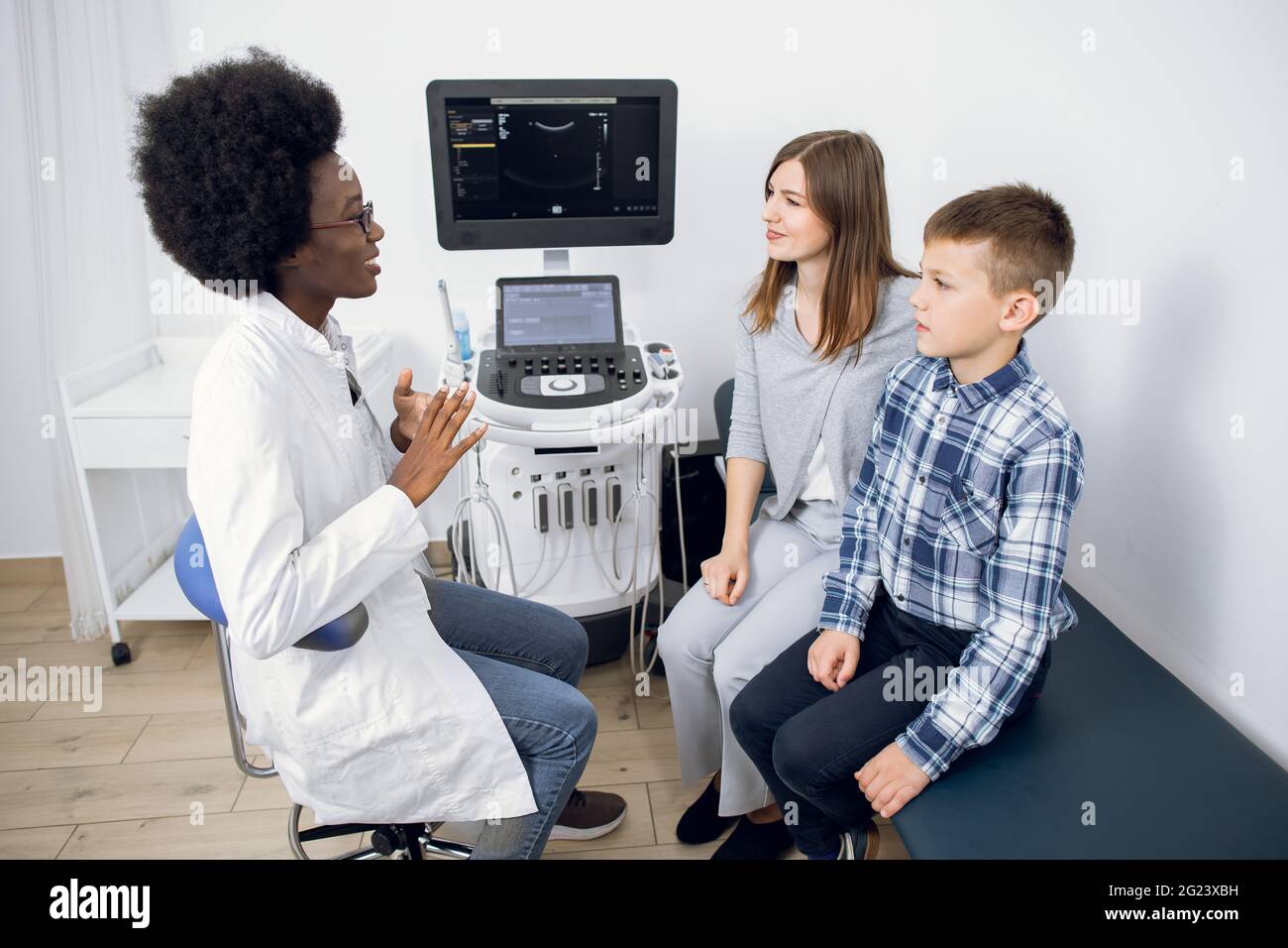 Junge kaukasische Frau im Alter von 30 Jahren mit ihrem Teenager-Sohn, die einen Arzt im modernen Ultraschalluntersuchungsraum besucht. afroamerikanische Ärztin erklärt ihren Patienten das Verfahren der Ultraschalluntersuchung Stockfoto