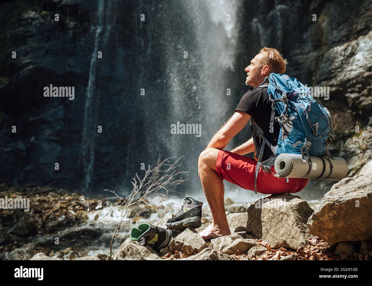 Mann mit Rucksack in aktiver Trekkingkleidung zieht Trekkingstiefel aus, sitzt in der Nähe des Gebirgswasserfalls und genießt die Natur. Reisen Stockfoto