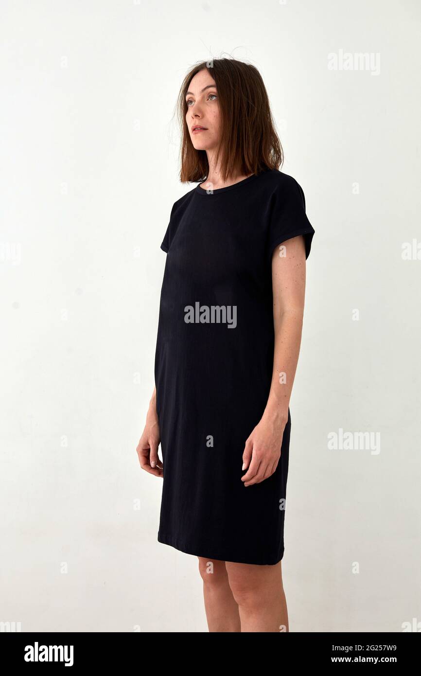 Erwachsenes weibliches Modell im lässigen einfachen schwarzen Midi-Kleid, das vor weißem Hintergrund steht und wegschaut Stockfoto