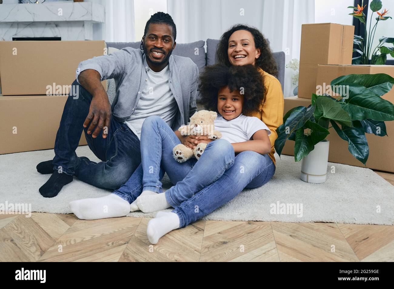 Glückliche gemischte Rennen Familie Spaß beim Umzug in eine neue Wohnung. Mutter, Vater und Tochter sitzen zusammen auf dem Boden eines neuen Hauses mit cardb Stockfoto