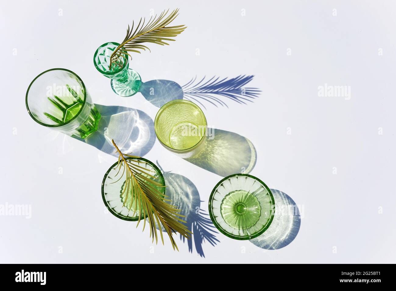 Sommerstill-Szene. Glitzernde grüne Gläser Wasser oder Cocktails auf weißem Tischhintergrund im Sonnenlicht. Lange harte Schatten. Urlaubskonzept Stockfoto