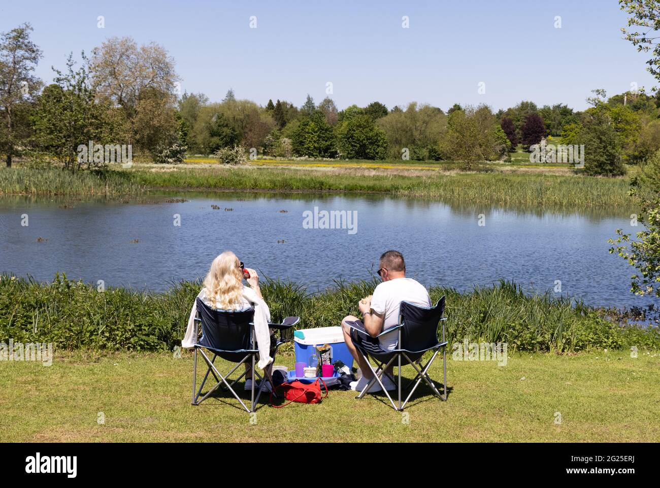 Kaukasisches Paar mittleren Alters, das auf Stühlen sitzt und an einem sonnigen Frühlingstag an einem See picknickt, Rückansicht, England Stockfoto
