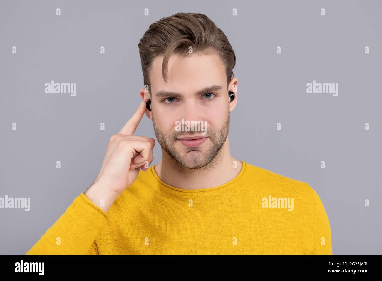 Schöner Kerl zeigen Finger auf Bluetooth-Ohrhörer im Ohr grauen Hintergrund, hören Stockfoto