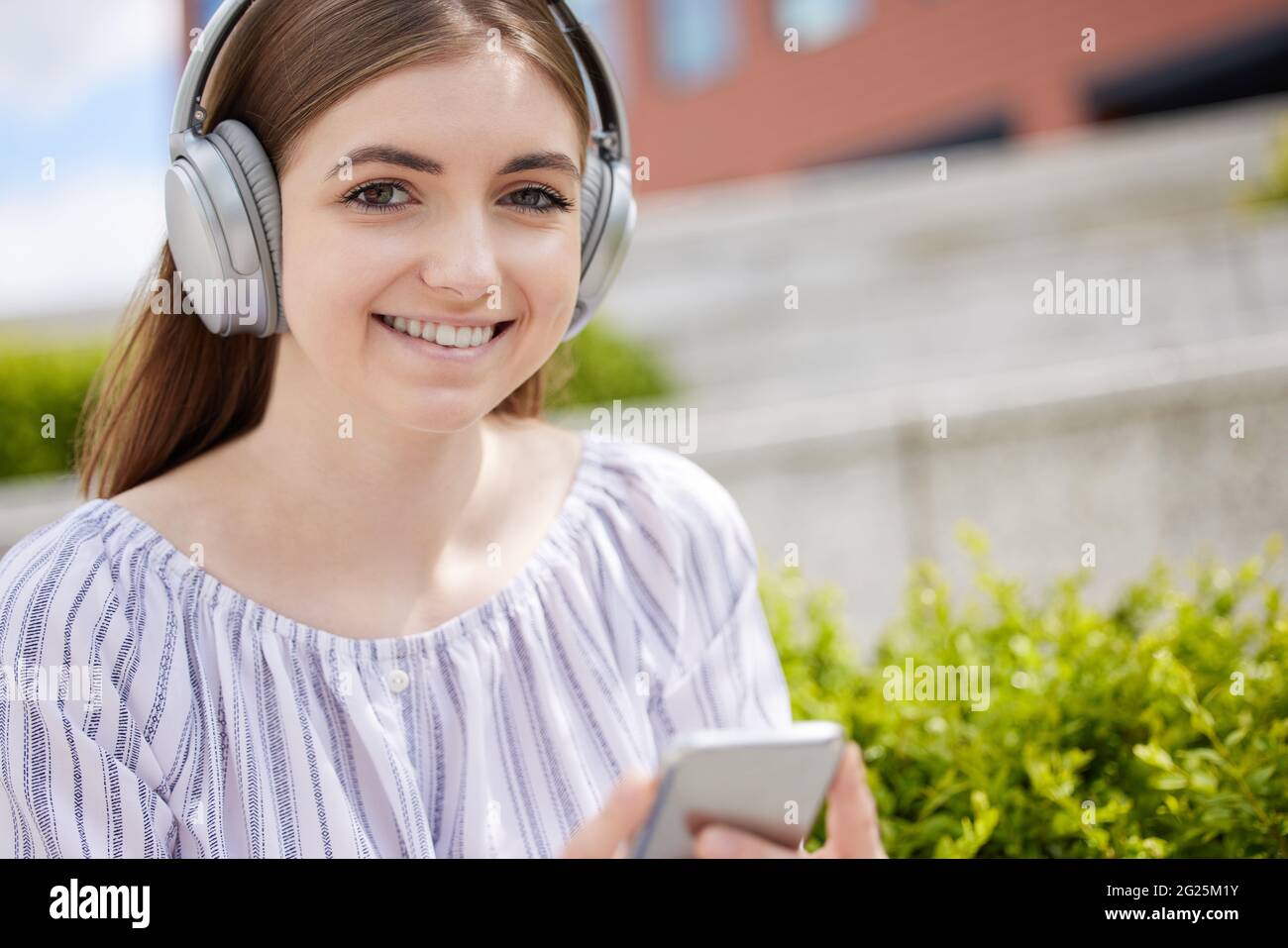 Portrait Einer Lächelnden Jungen Studentin Auf Dem College Campus Streamt Musik Vom Mobiltelefon Auf Kabellose Kopfhörer Stockfoto