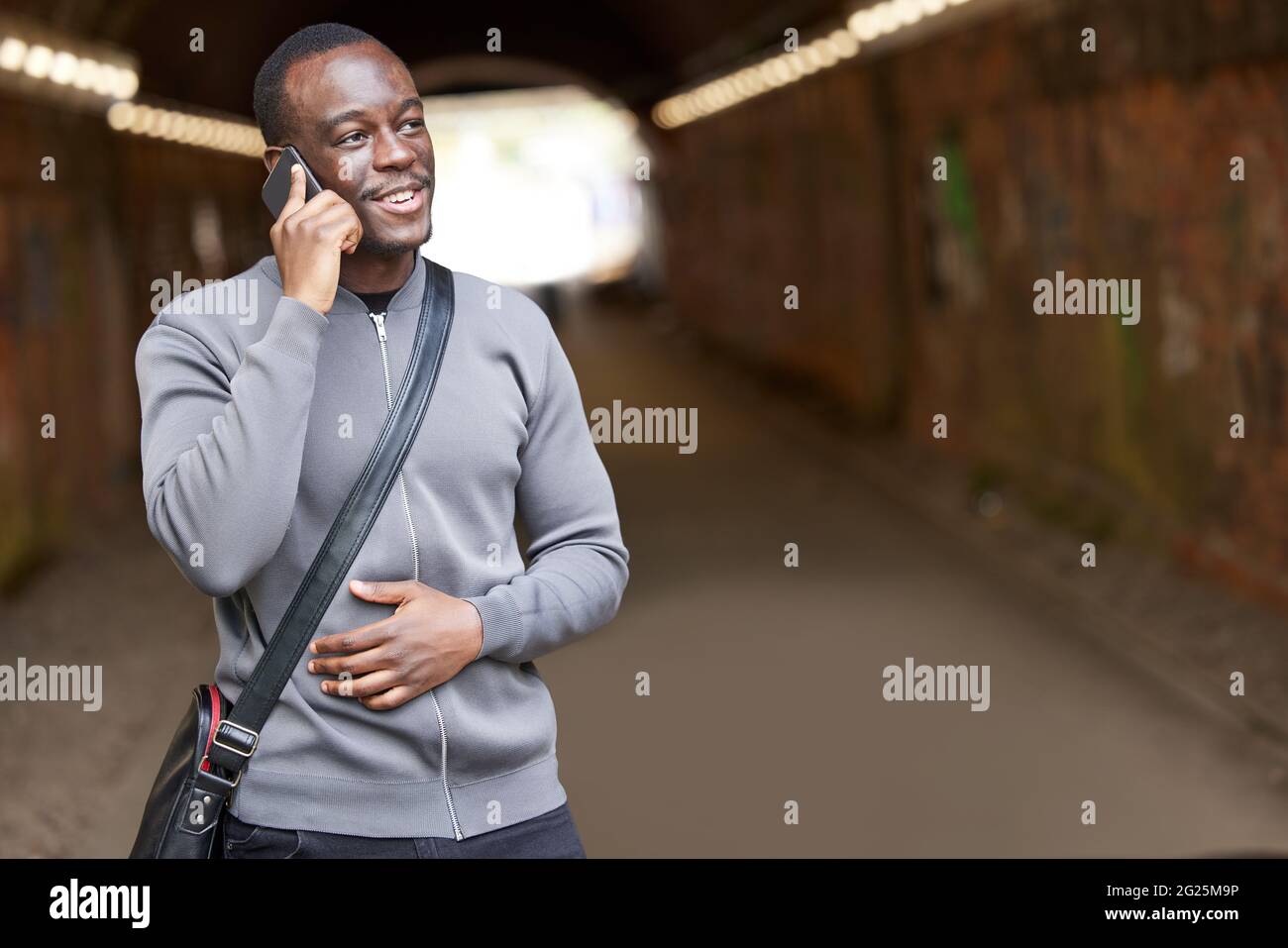 Lächelnder Junger Mann In Städtischer Umgebung Steht Im Tunnel Und Spricht Auf Dem Mobiltelefon Stockfoto