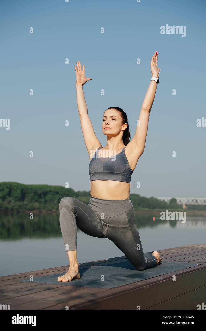 Selbstgefällige junge Frau, die im Freien an einem schönen Ort auf einer Seite des Flusses Yoga macht. Bäume spiegeln sich im Wasser. An einem sonnigen Tag unter einer klaren blu Stockfoto