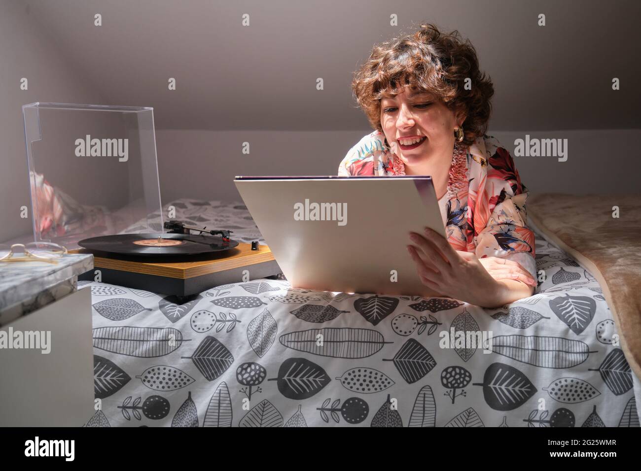 Junge Frau, die auf ihrem Bett lag, eine Schallplatte hörte und auf den Ärmel schaute Stockfoto