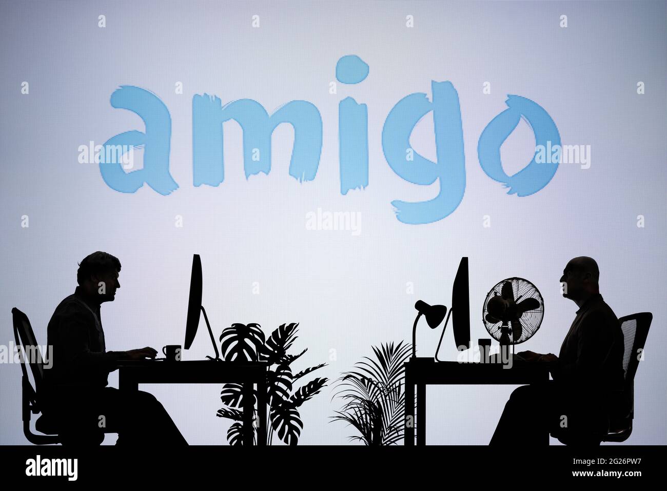 Das Logo der Amigo-Darlehen ist auf einem LED-Bildschirm im Hintergrund zu sehen, während zwei silhouettierte Menschen in einer Büroumgebung arbeiten (nur redaktionelle Verwendung) Stockfoto