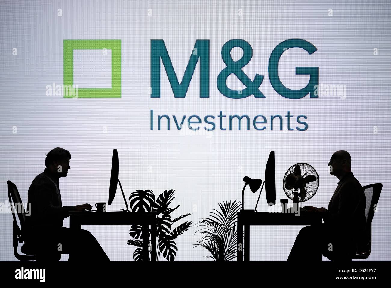 Das M&G Investments-Logo wird auf einem LED-Bildschirm im Hintergrund angezeigt, während zwei silhouettierte Personen in einer Büroumgebung arbeiten (nur zur redaktionellen Verwendung) Stockfoto