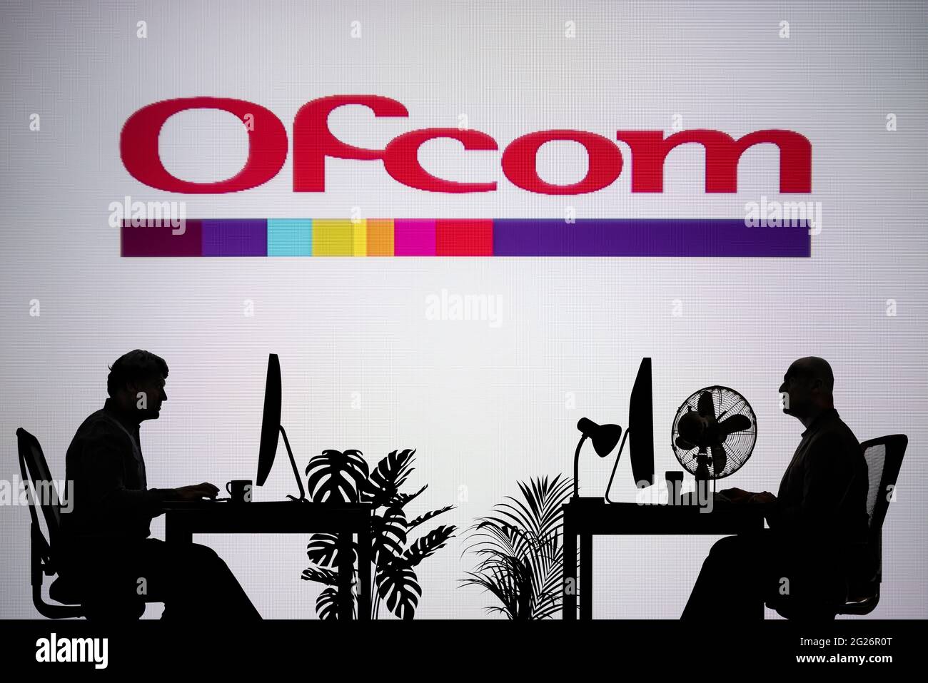 Das Ofcom-Logo ist auf einem LED-Bildschirm im Hintergrund zu sehen, während zwei silhouettierte Menschen in einer Büroumgebung arbeiten (nur zur redaktionellen Verwendung) Stockfoto