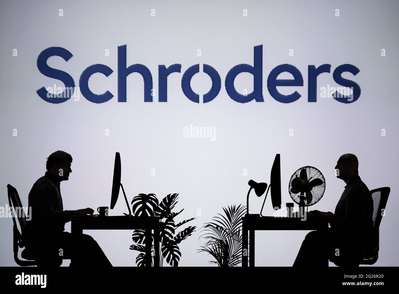 Das Schroders-Logo ist auf einem LED-Bildschirm im Hintergrund zu sehen, während zwei silhouettierte Personen in einer Büroumgebung arbeiten (nur zur redaktionellen Verwendung) Stockfoto