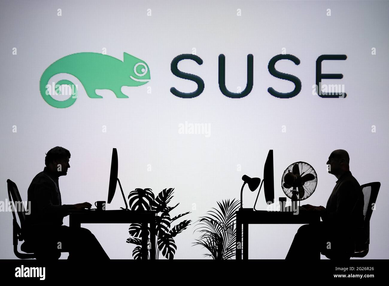 Das SUSE Logo wird auf einem LED-Bildschirm im Hintergrund angezeigt, während zwei silhouettierte Personen in einer Büroumgebung arbeiten (nur für redaktionelle Zwecke). Stockfoto