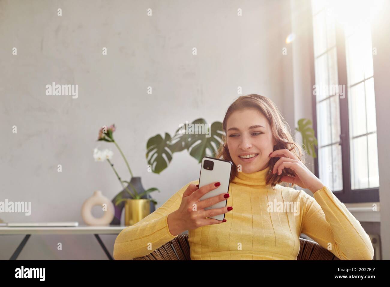 Glückliche Frau in gelbem Rollkragen winkende Geste, während sie auf einem Stuhl sitzt und auf einem Mobiltelefon in einem hellen Arbeitsbereich schaut Stockfoto