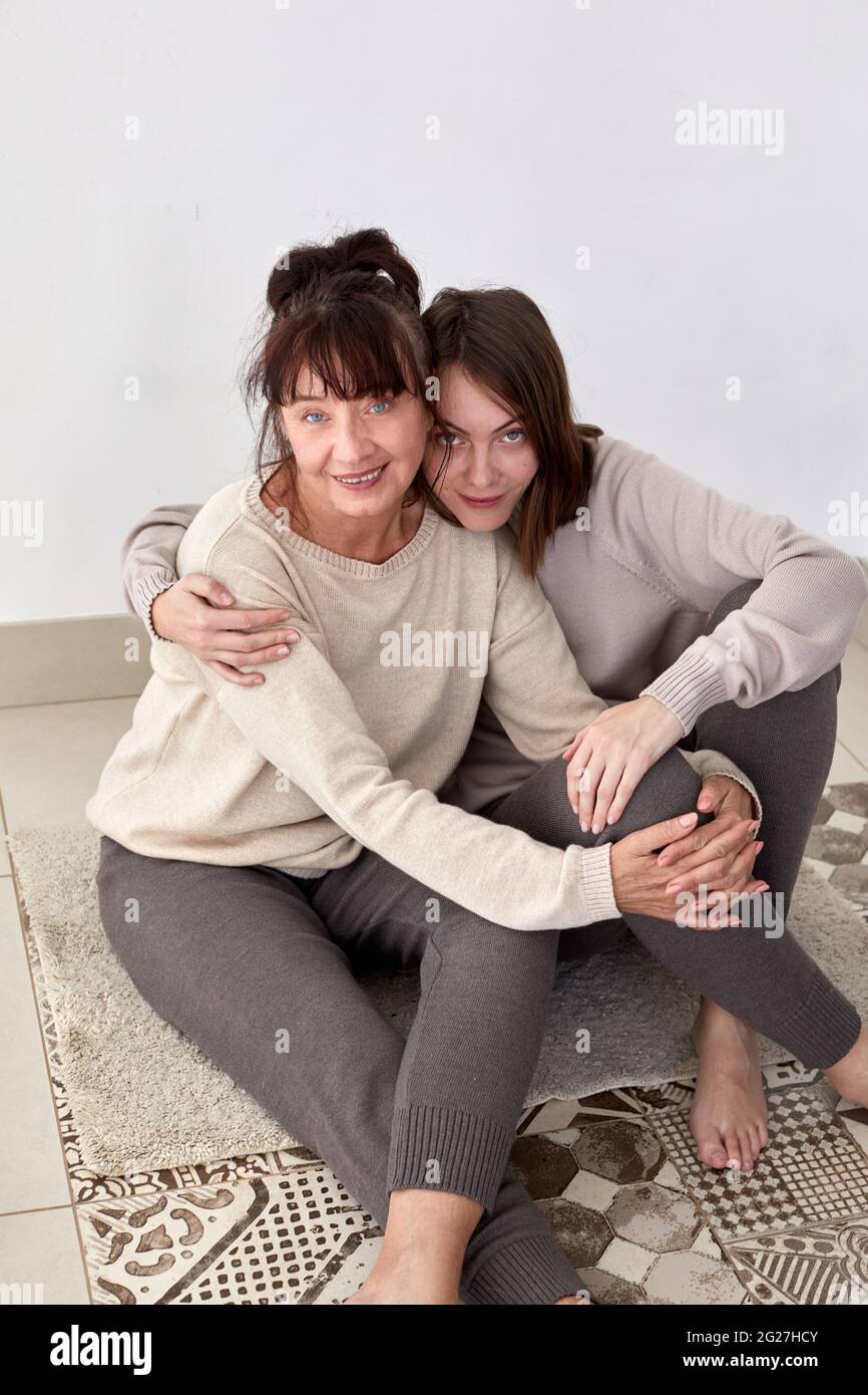 Ein hoher Blickwinkel von glücklicher junger Frau und Mutter mittleren Alters in bequemen, lässigen Outfits, die die Kamera umarmen und betrachten Stockfoto Ein hoher Blickwinkel von glücklicher junger Frau und Mutter mittleren Alters in bequemen, lässigen Outfits, die die Kamera umarmen und betrachten Stockfoto