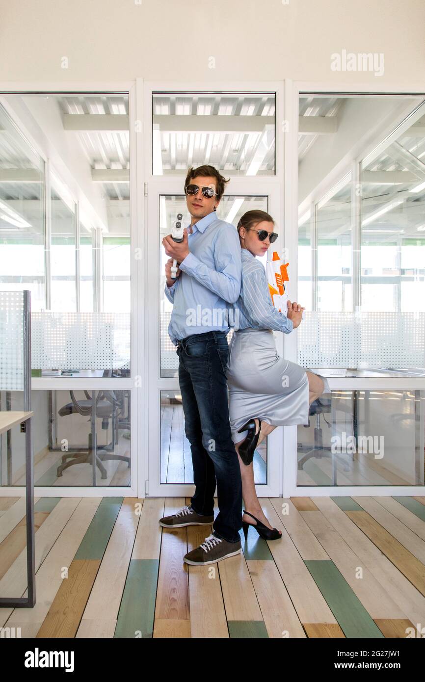 Ganzkörperansicht von Büromitarbeitern in Sonnenbrillen, die am Arbeitsplatz mit Spielzeugpistolen Rücken an Rücken stehen Stockfoto