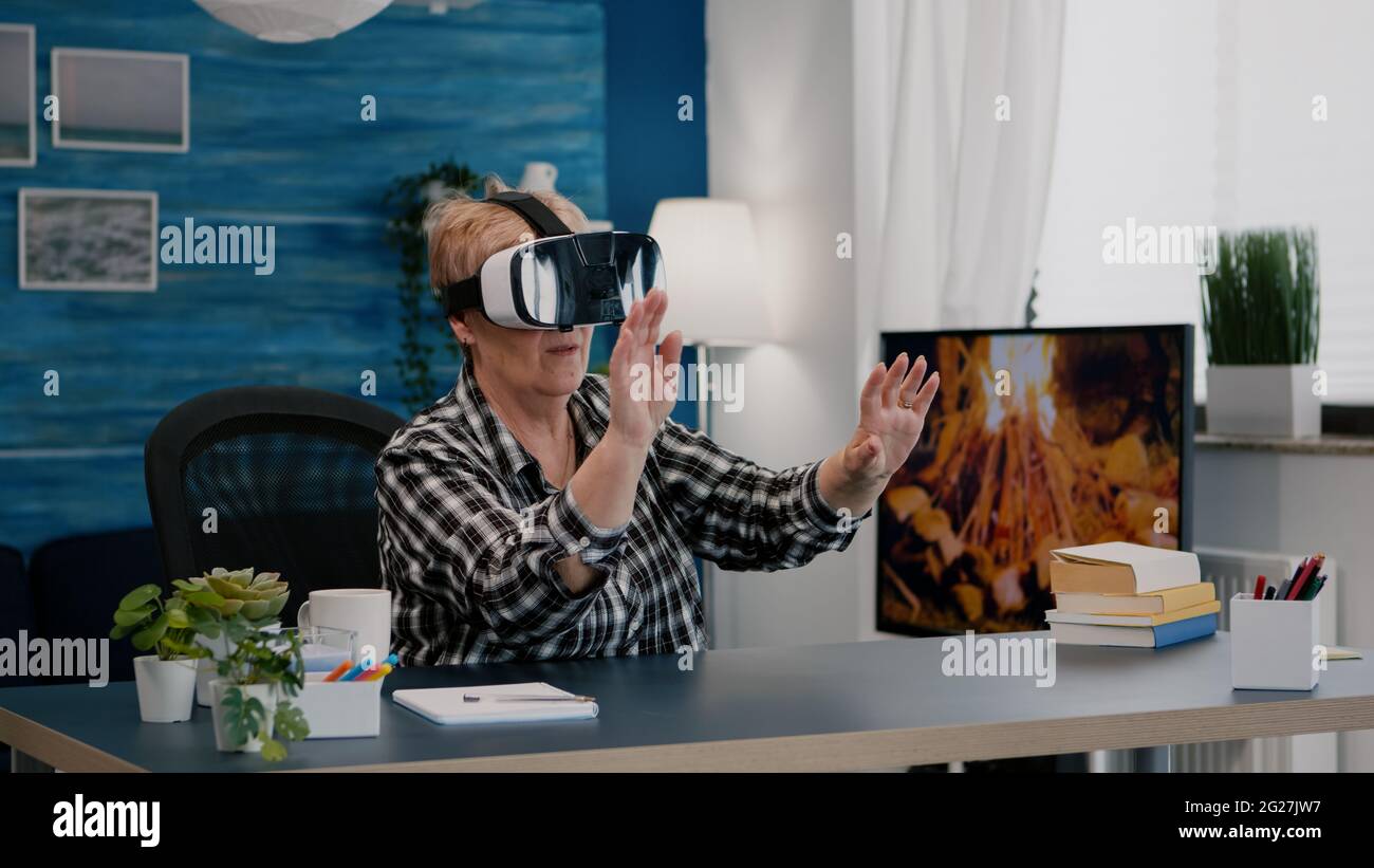 Pensionierte Frau erlebt virtuelle Realität mit vr-Headset im Wohnzimmer arbeiten von zu Hause aus. Alte Remote-Mitarbeiter suchen, Finanzberichte analysieren, während ältere Frau im Hintergrund Fernsehen schaut Stockfoto