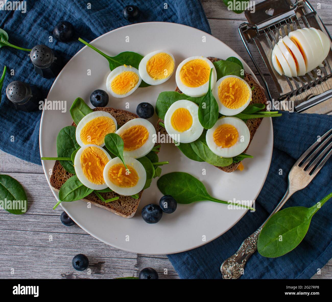 Eiersandwich aus Roggenbrot und gekrönt mit gekochtem und geschnittenem Ei und Spinat. Serviert mit frischen Blaubeeren auf einem Teller auf einem Holztisch Stockfoto