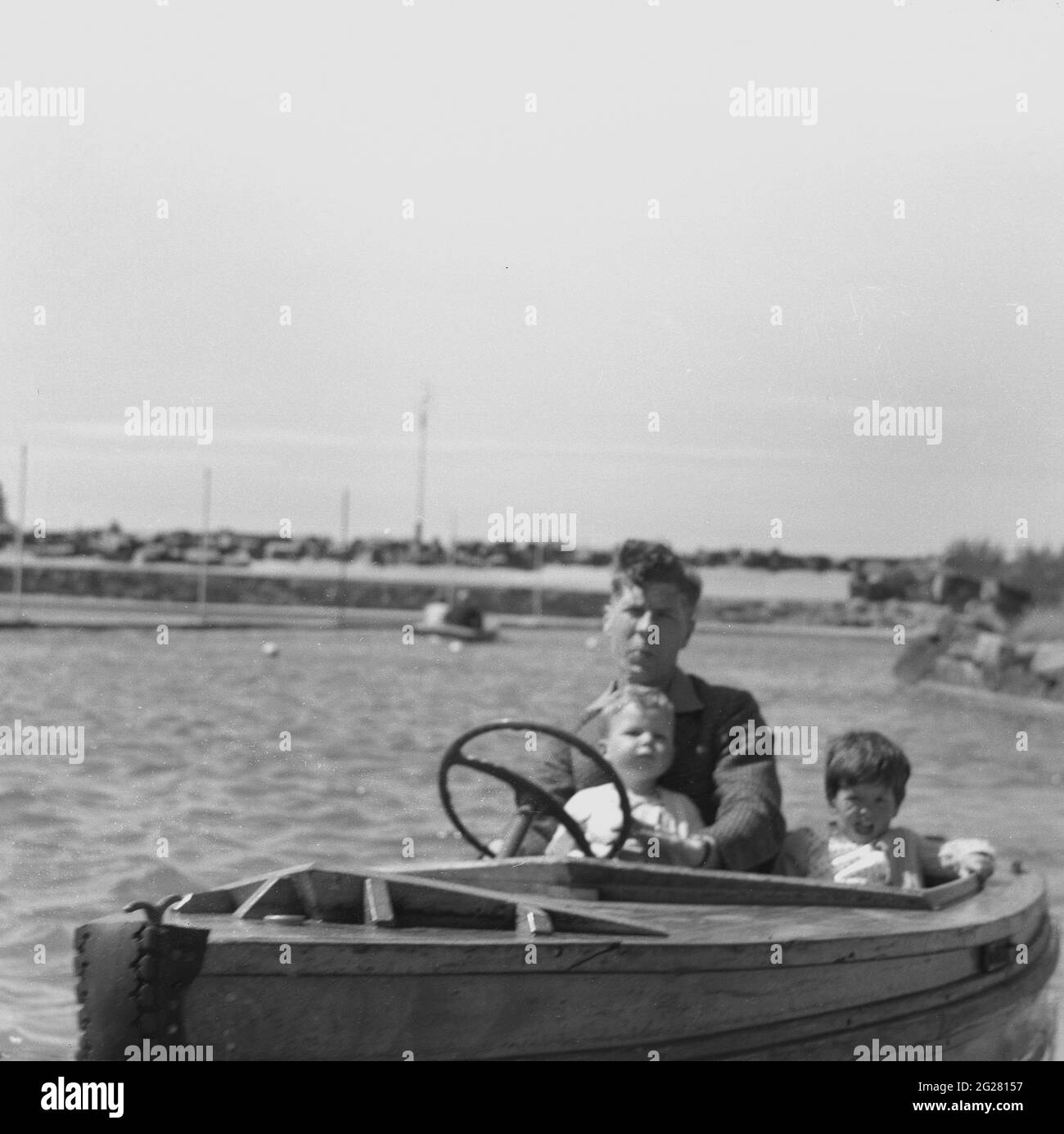 In den 1950er Jahren, ein Vater mit seinen zwei Kleinkindern, der ein kleines Holzboot auf einem See mit Bootstouren am Meer steuerte, England, Großbritannien. Stockfoto