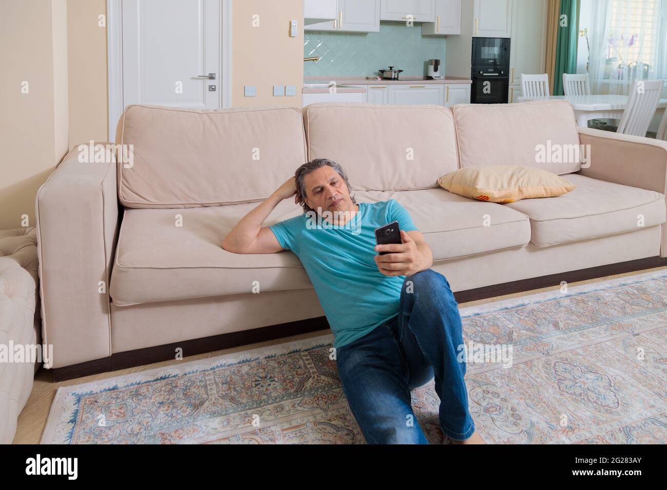 Ein erwachsener Mann durchsucht soziale Netzwerke auf einem Mobiltelefon, während er in einer Wohnung auf dem Boden sitzt. Stockfoto